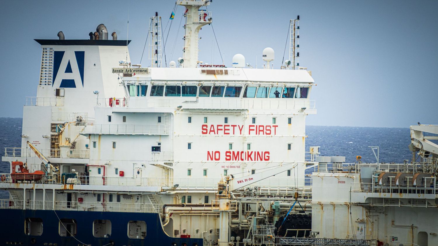Large white cargo ship with red Safety First No Smoking sign on hull, moored at sea under cloudy sky.