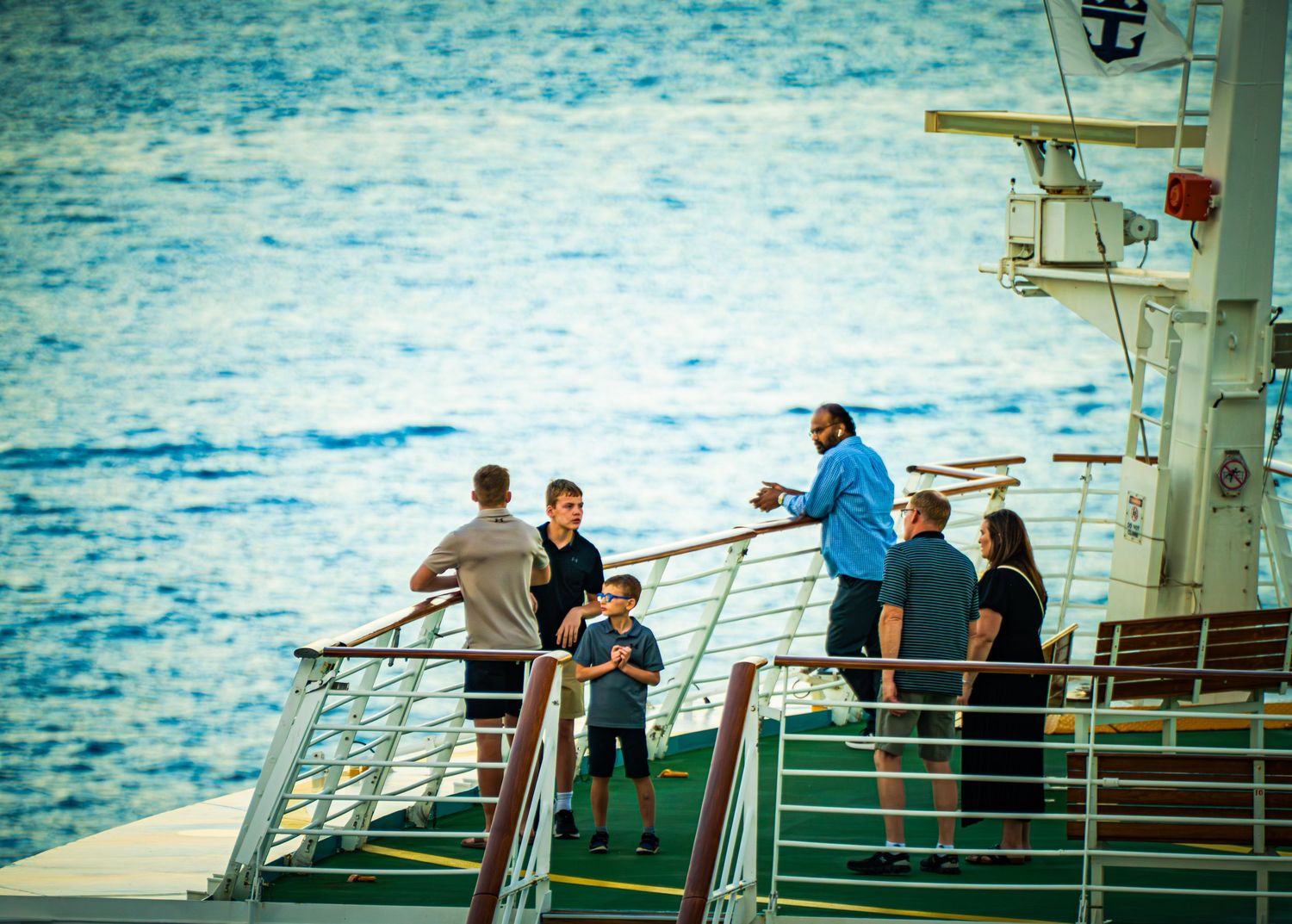 Passengers gather on the deck of a cruise ship against a bright blue ocean backdrop.