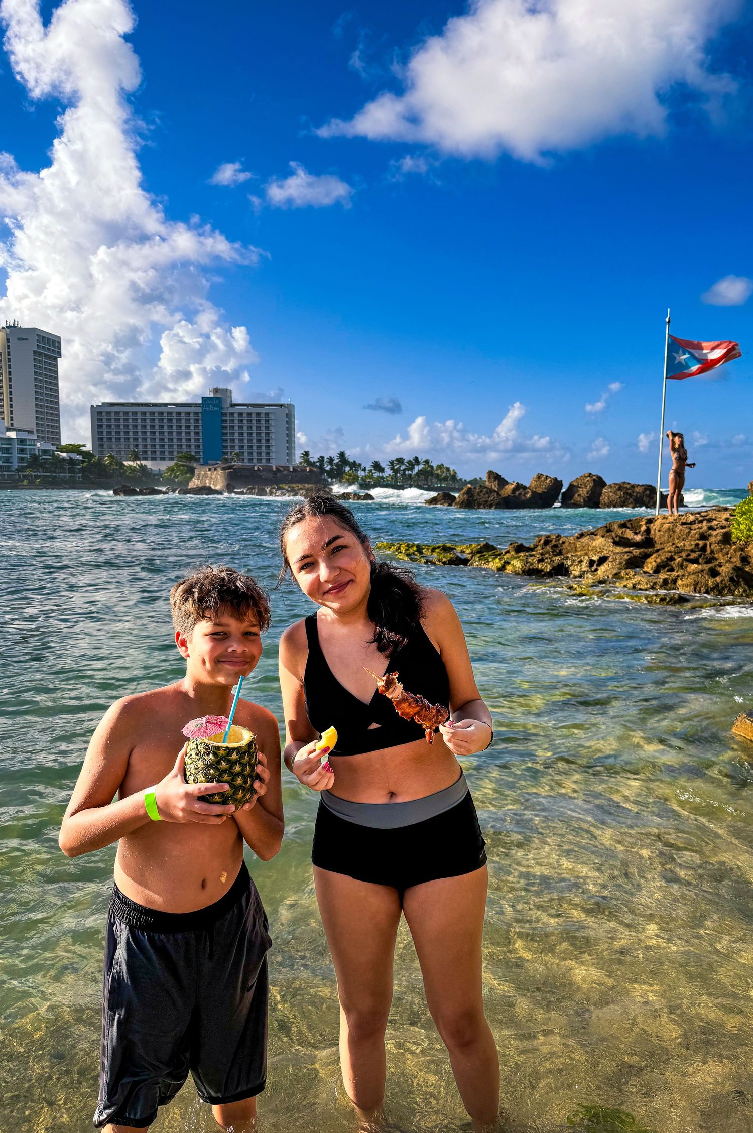 People enjoying a sunny day at a tropical beach with clear blue water and resort buildings in the background.