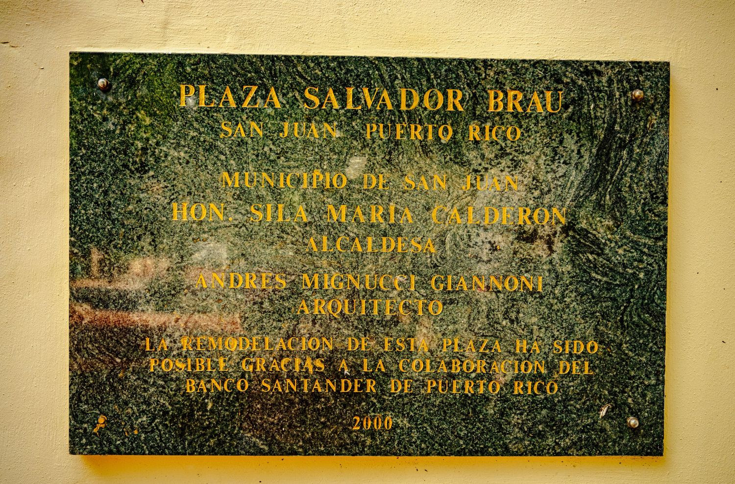 A weathered brass memorial plaque with engraved text mounted on a textured green surface.