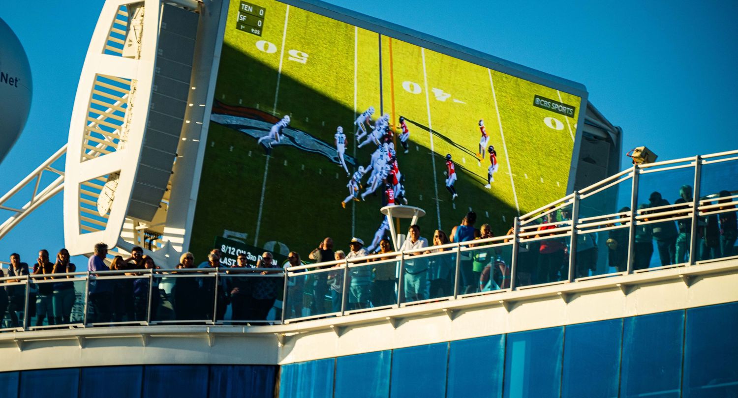 Crowds gather on a modern bridge walkway with bright blue and green architectural elements against a clear sky.