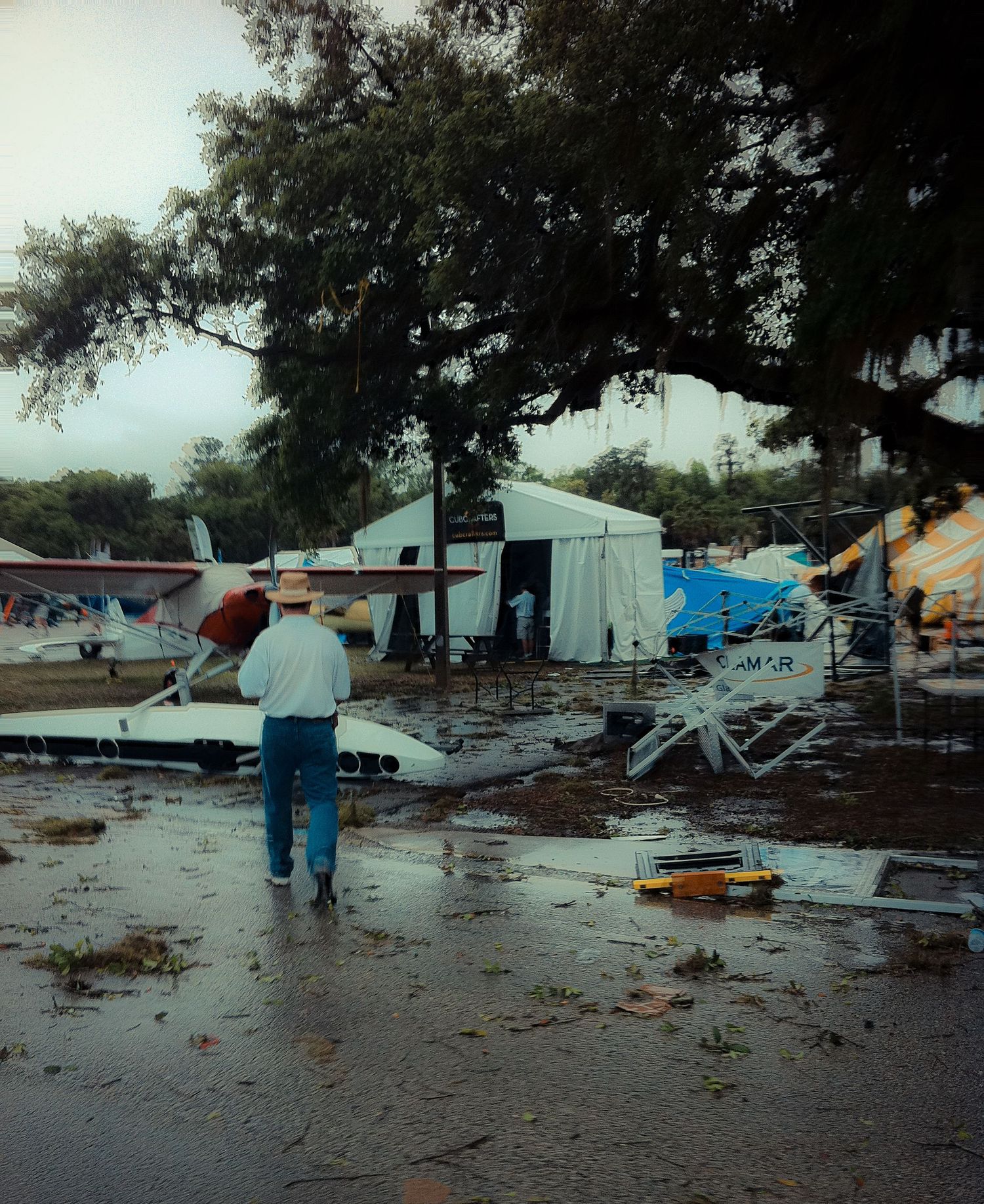Aftermath of storm damage at a campground showing debris and wet ground with tents and temporary structures in background.