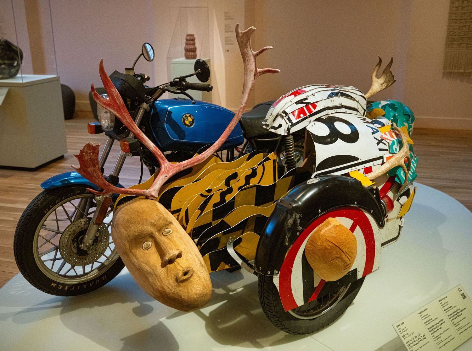 Colorful custom motorcycle art installation with decorative masks and painted designs on display in gallery setting.