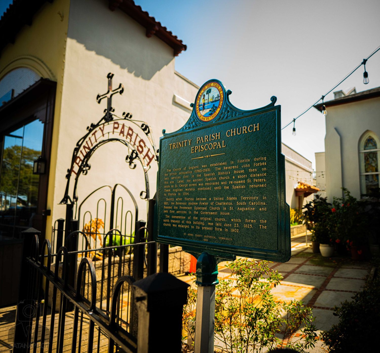 Ornate iron gates and historical marker signs frame the entrance to a historic church under golden sunlight.