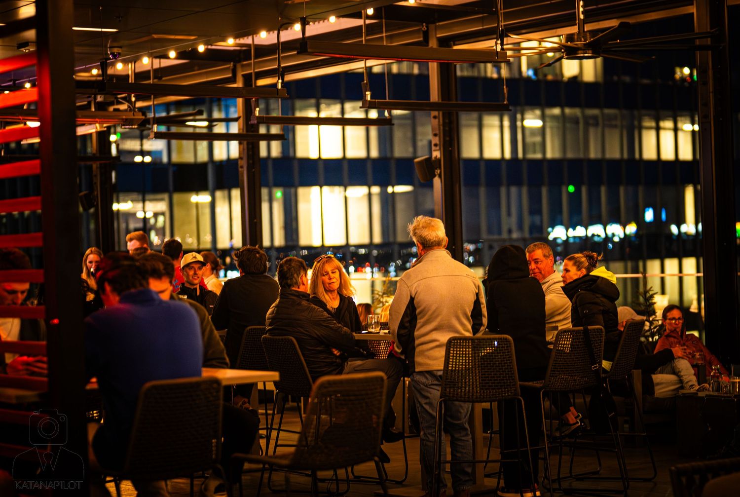 Nighttime diners enjoy outdoor seating at an urban restaurant with illuminated office buildings in the background.