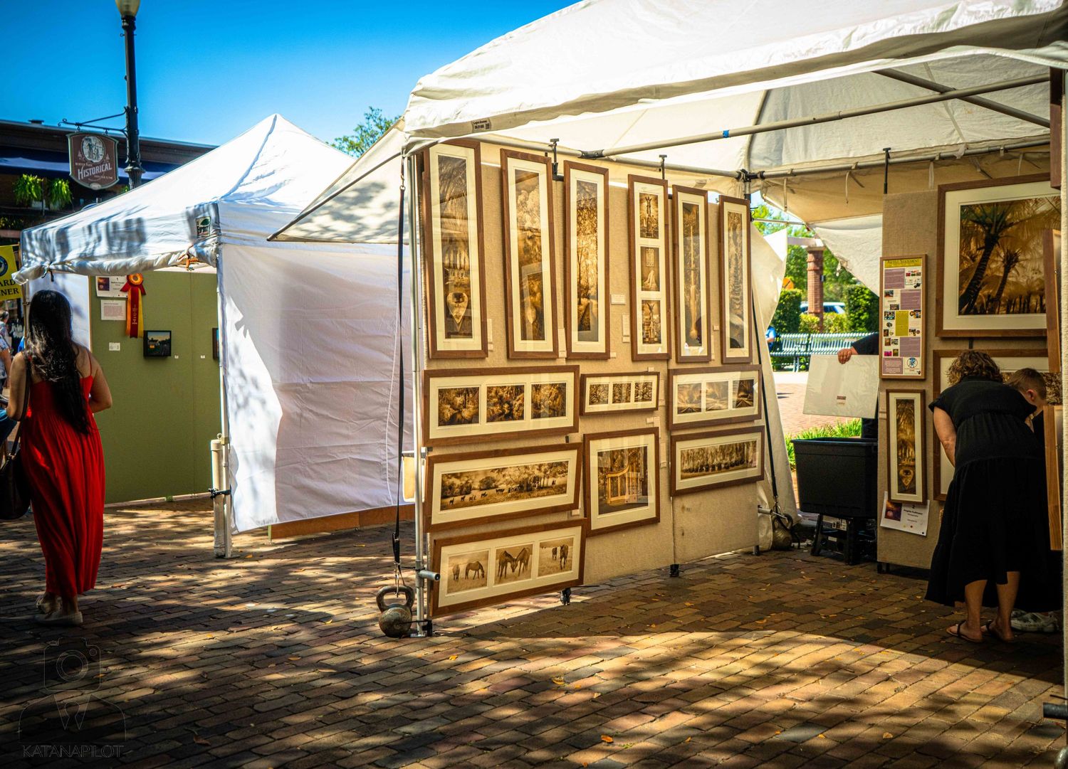 Art vendor displays framed artwork at an outdoor festival market with white tents and dappled sunlight.