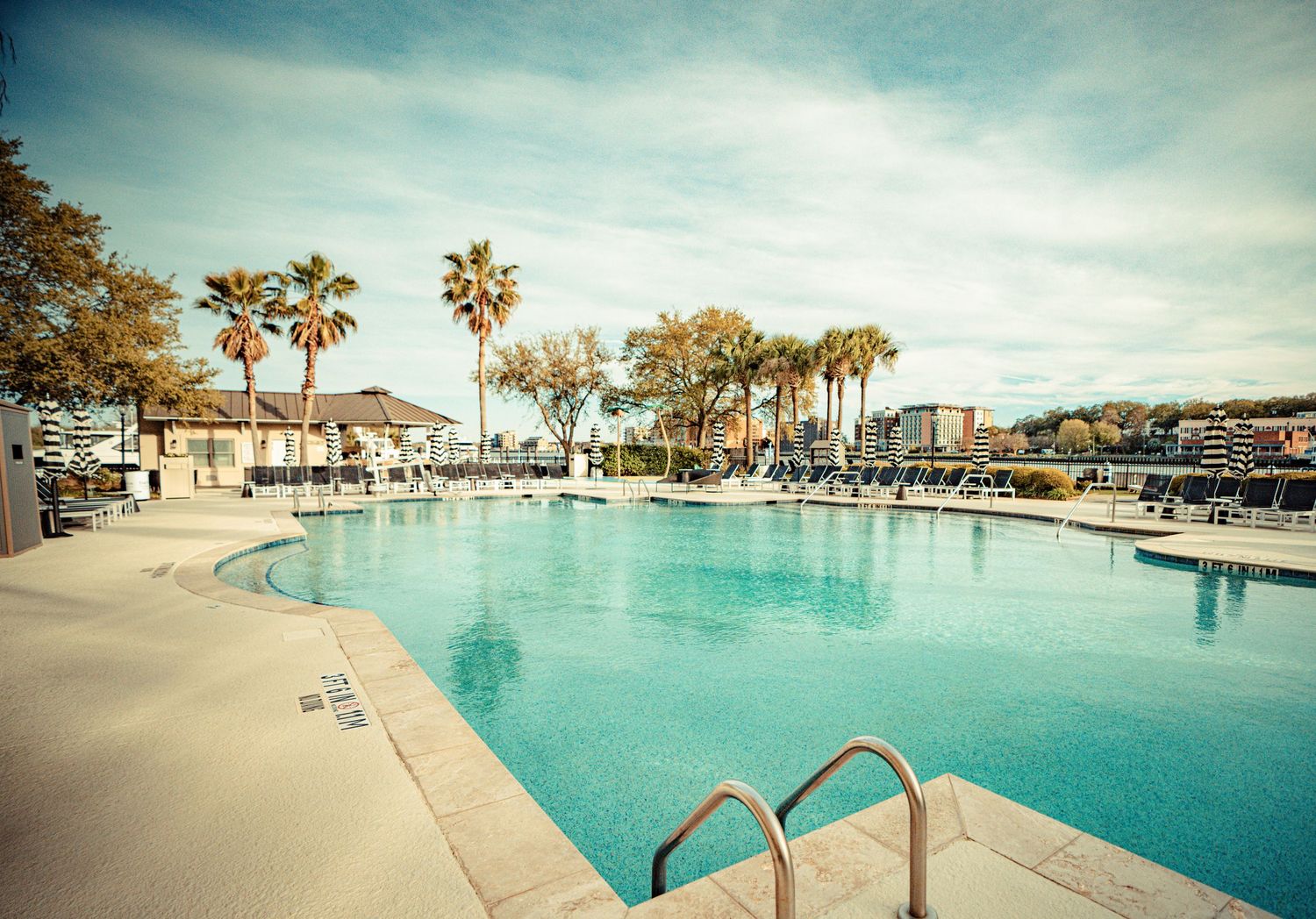 Panoramic view of a resort swimming pool with palm trees and lounge chairs under a bright turquoise sky.