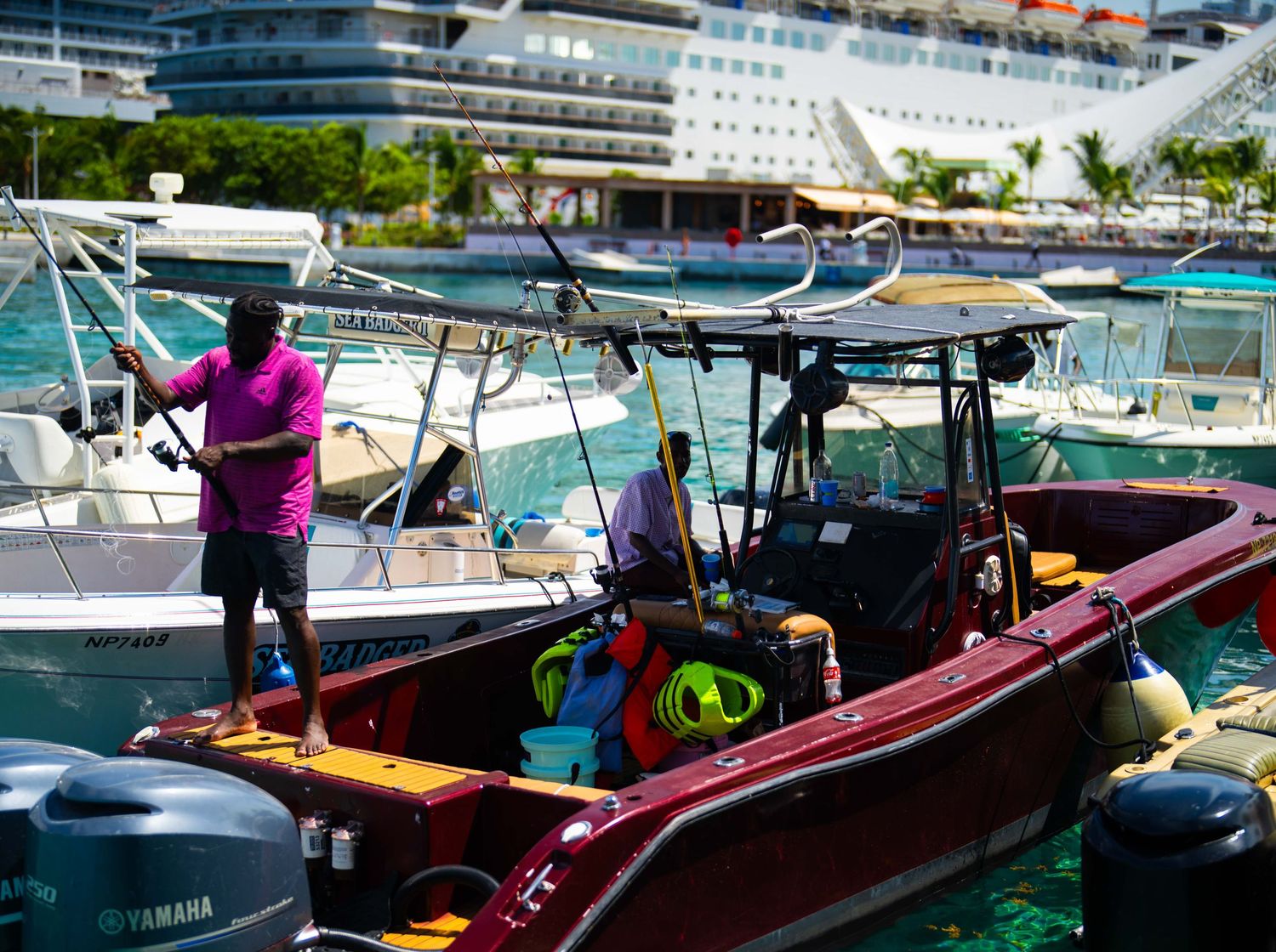 Two men working on a red motorboat docked in a tropical harbor with a cruise ship in the background.