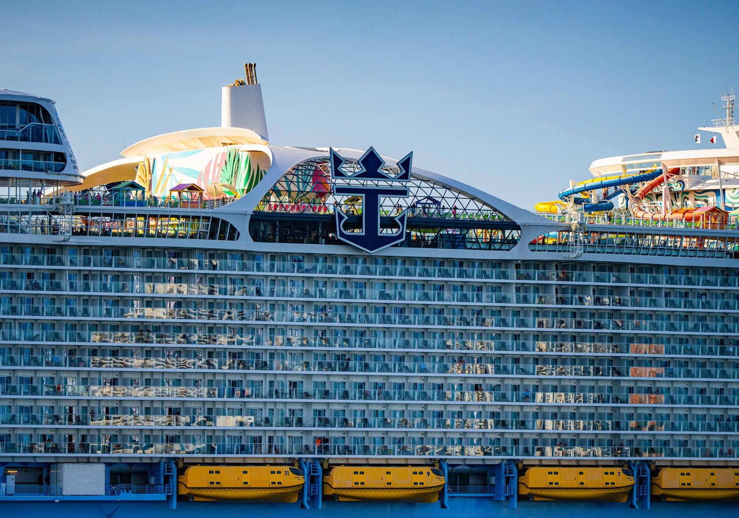 Large Royal Caribbean cruise ship docked at port, featuring colorful water slides and multiple decks of balconies under clear blue sky.
