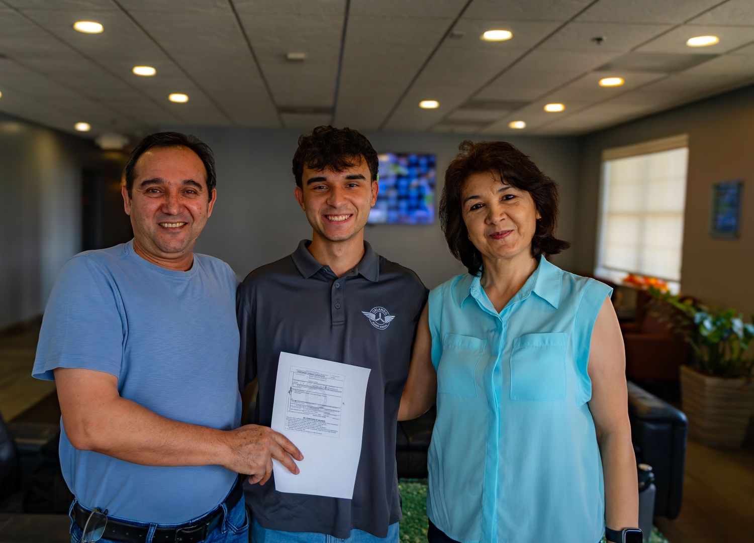 Family of three proudly holding a certificate together, smiling indoors at a special achievement ceremony.