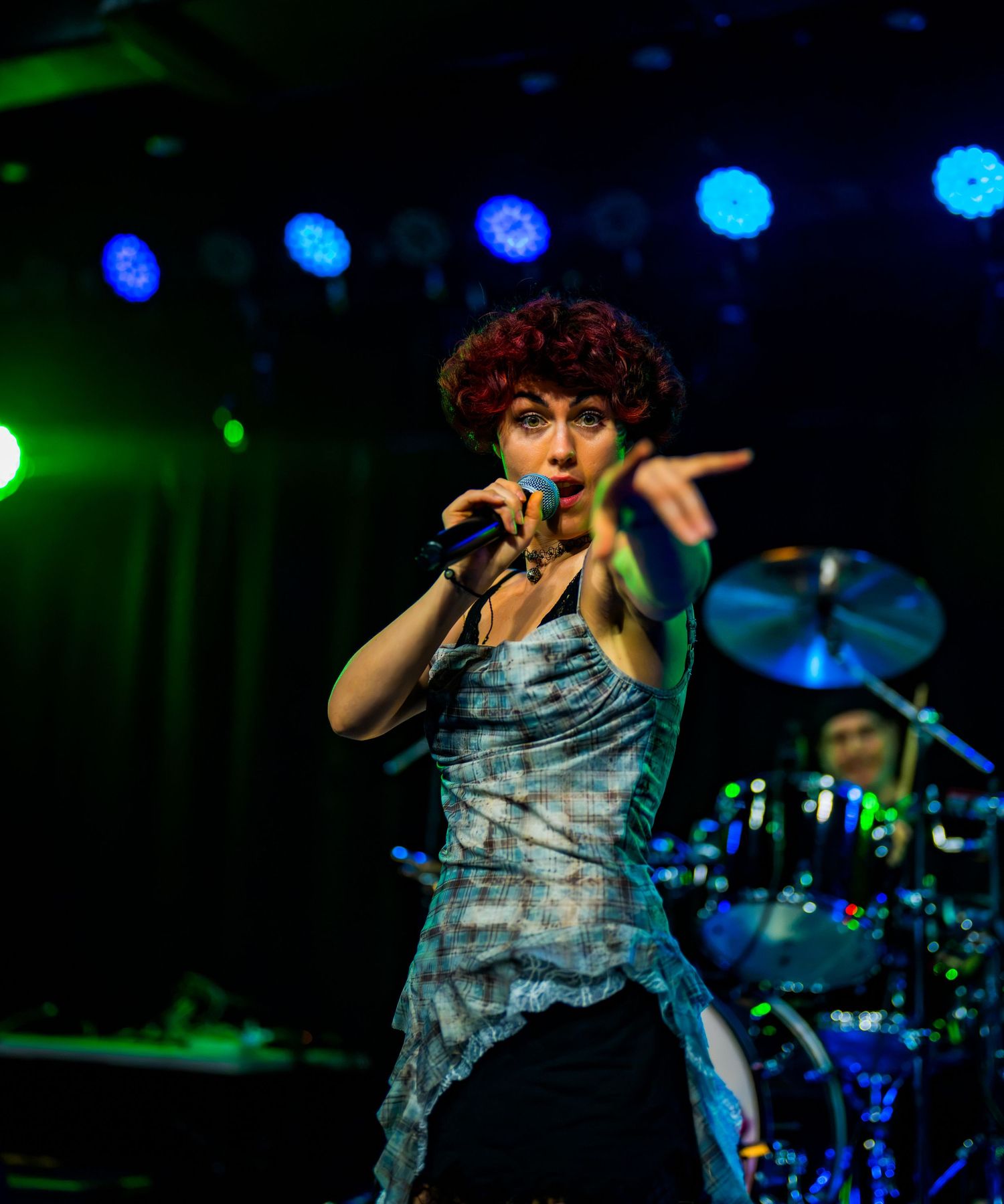 Red-haired female singer performing on stage with blue lights and drummer in background.