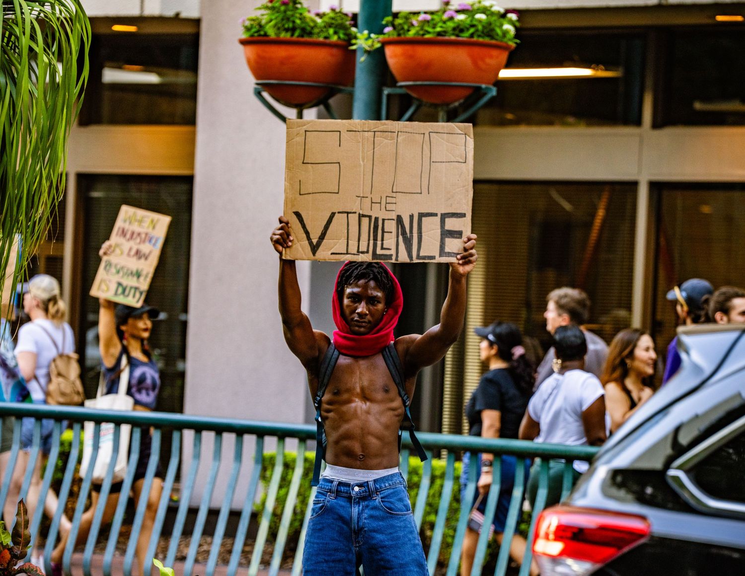 Shirtless protester holds sign during street demonstration with planters visible in background.