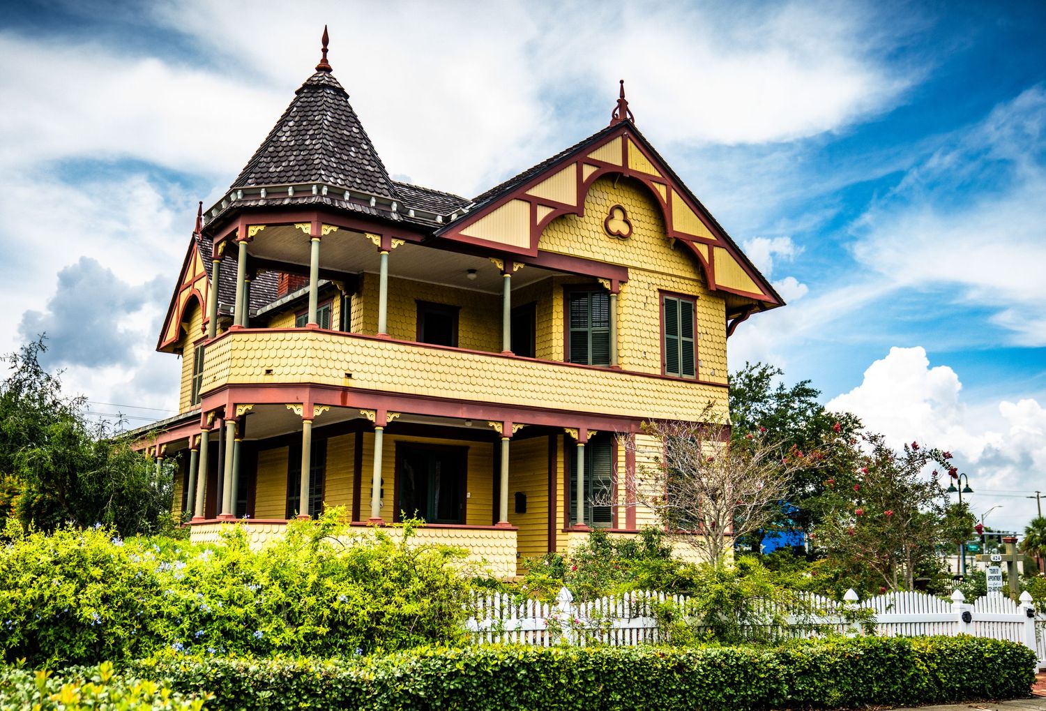 Victorian yellow mansion with ornate details and wraparound porch against blue sky.