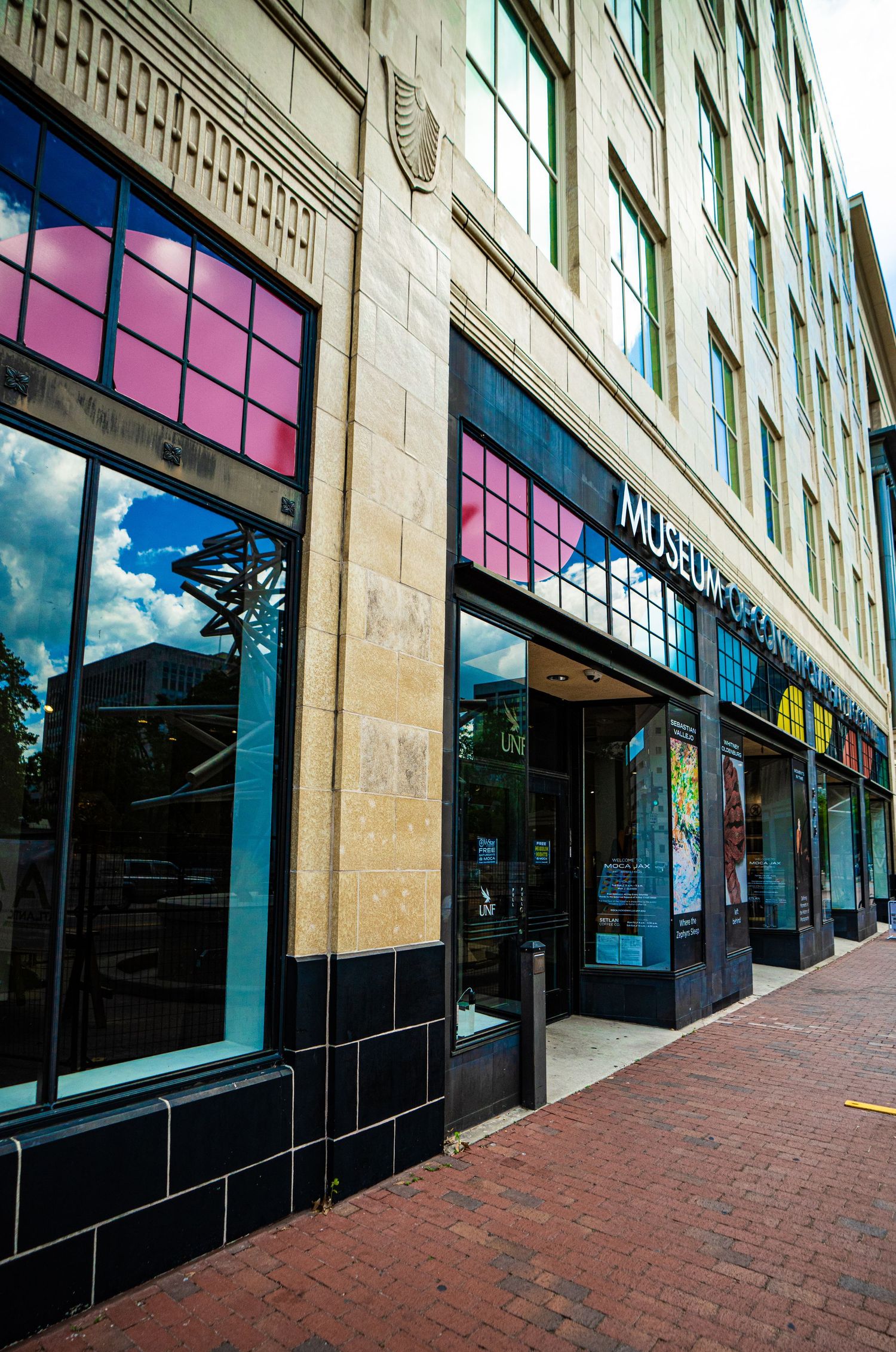 Colorful museum entrance with pink and blue windows on a brick sidewalk in an urban downtown building.
