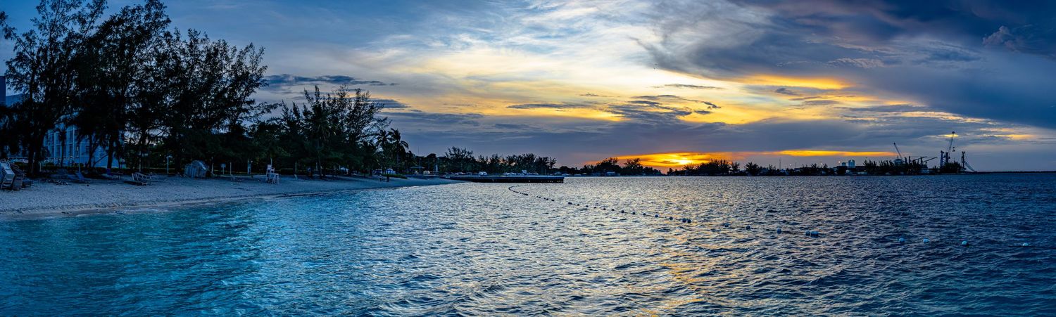 Stunning panoramic sunset over a tropical beach with vibrant blue waters and silhouetted palm trees on the horizon.