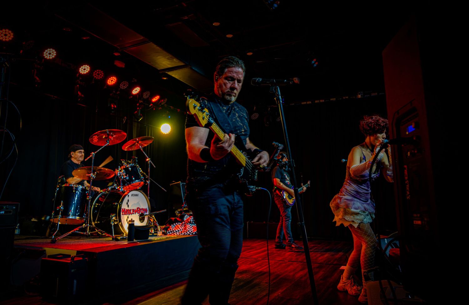 Rock band performing on stage with guitarist, drummer, and female vocalist under dramatic red and blue stage lighting.