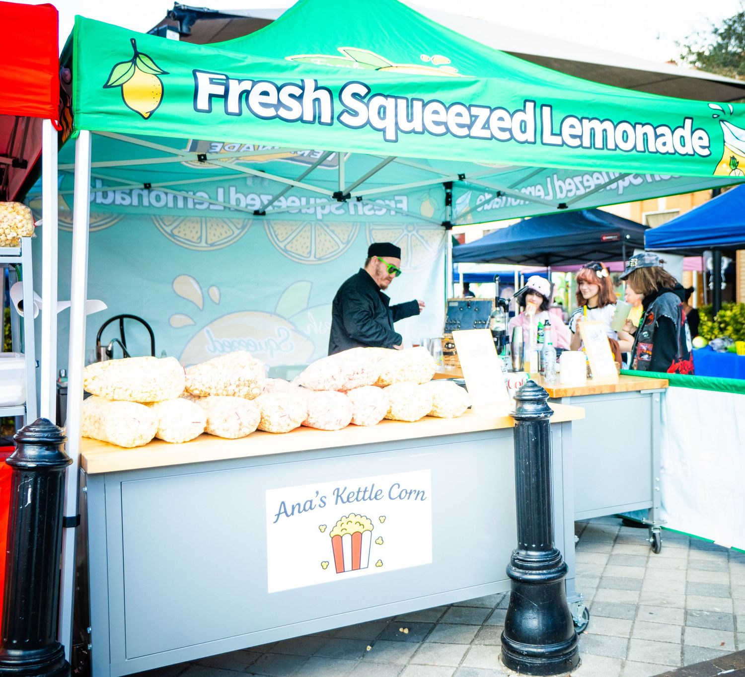 A vendor sells fresh squeezed lemonade from a turquoise tent at an outdoor market with stacks of cups on display.