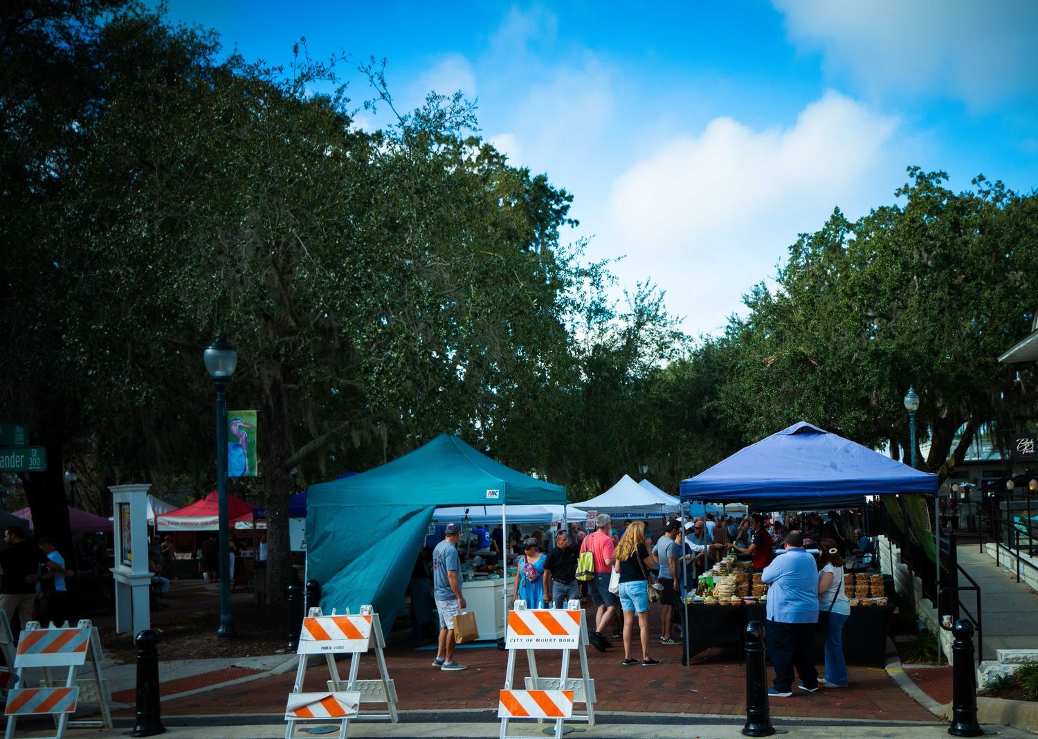Outdoor market stalls with blue tents set up along a tree-lined street on a sunny day.