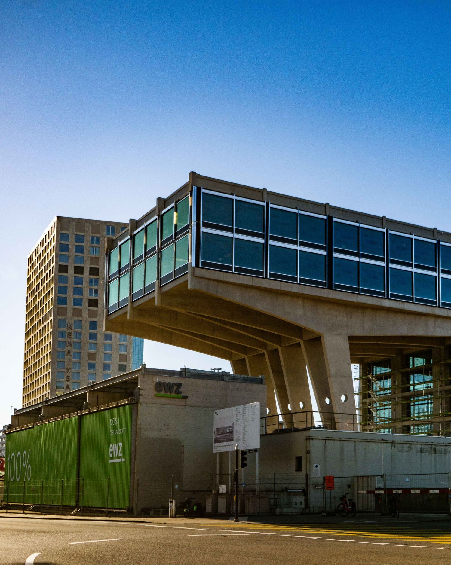Modern glass-walled building on massive concrete supports above a green industrial structure on a city street under a clear blue sky.