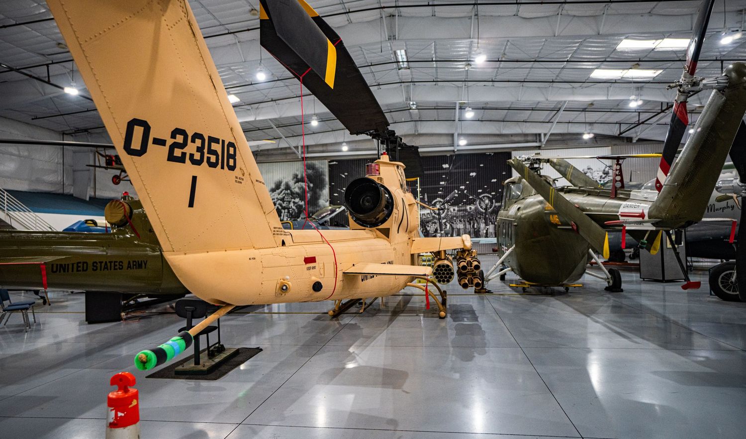 Military helicopters on display inside a spacious aviation museum hangar, including a tan U.S. Army helicopter marked D-23518.