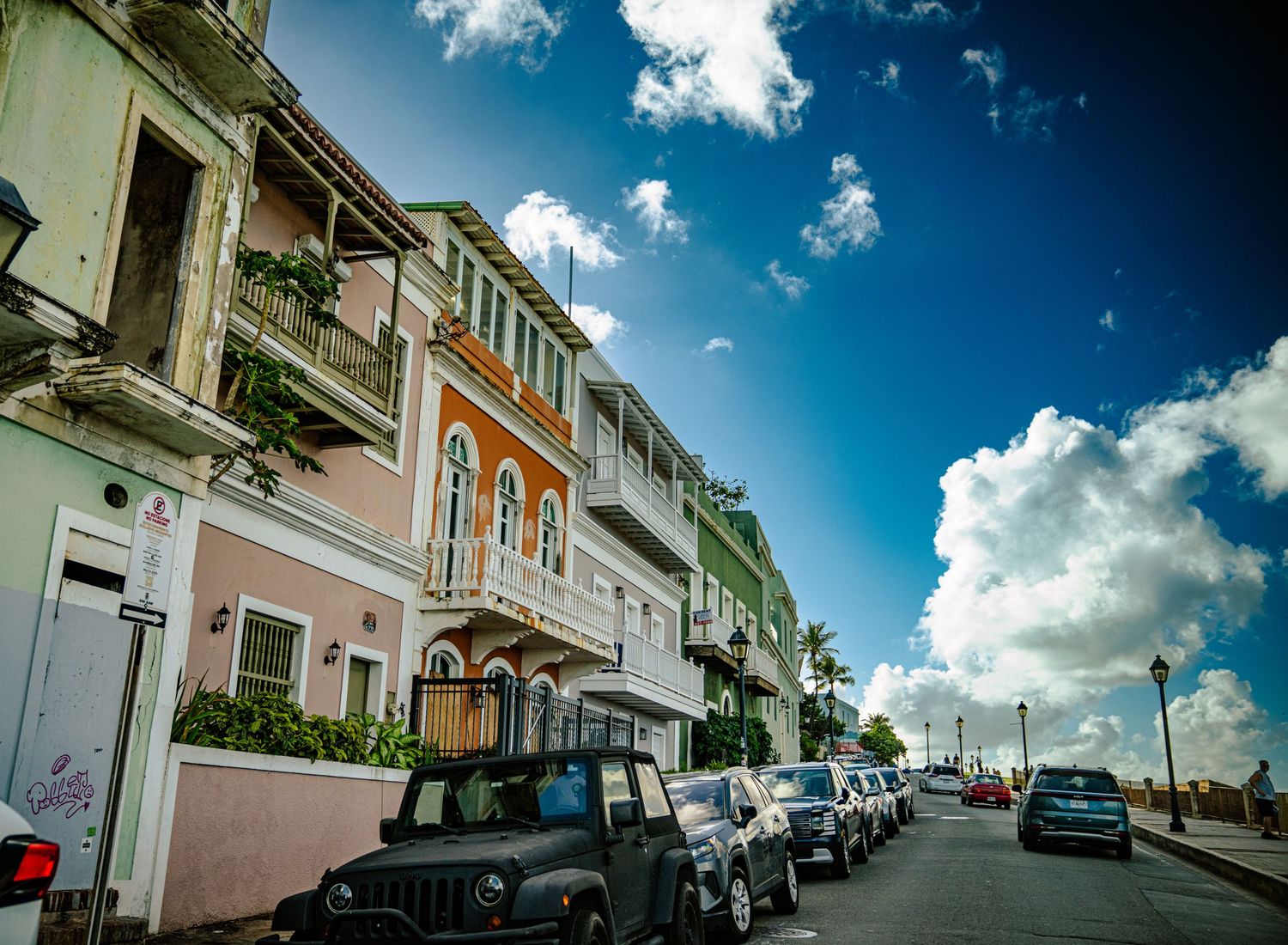 A panoramic view of a colorful colonial street with parked cars and white puffy clouds in a bright blue sky.