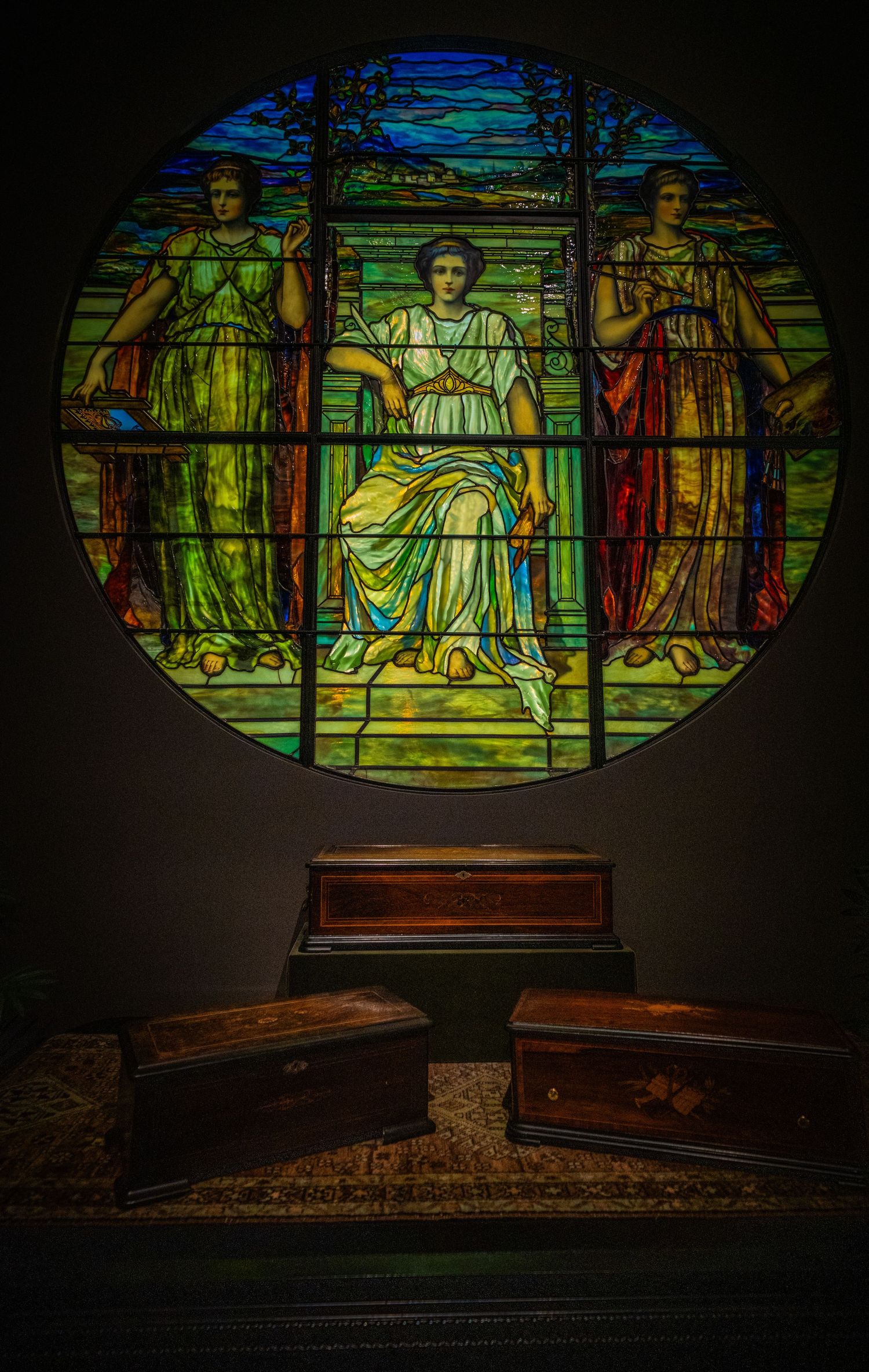 Round stained glass window above wooden chests depicting three robed figures in vibrant colors.