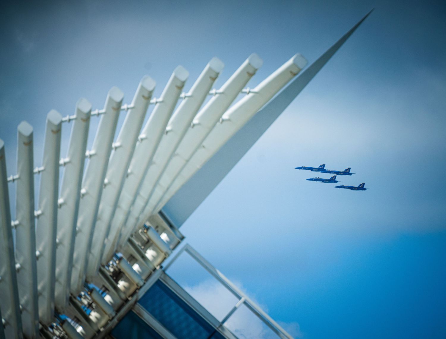 Modern architectural structure with white angular beams against blue sky as aircraft flies overhead.