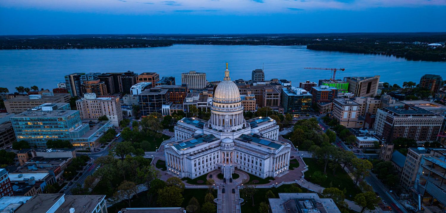 Aerial dusk view of Wisconsin State Capitol building with golden dome surrounded by Madison cityscape and Lake Monona.