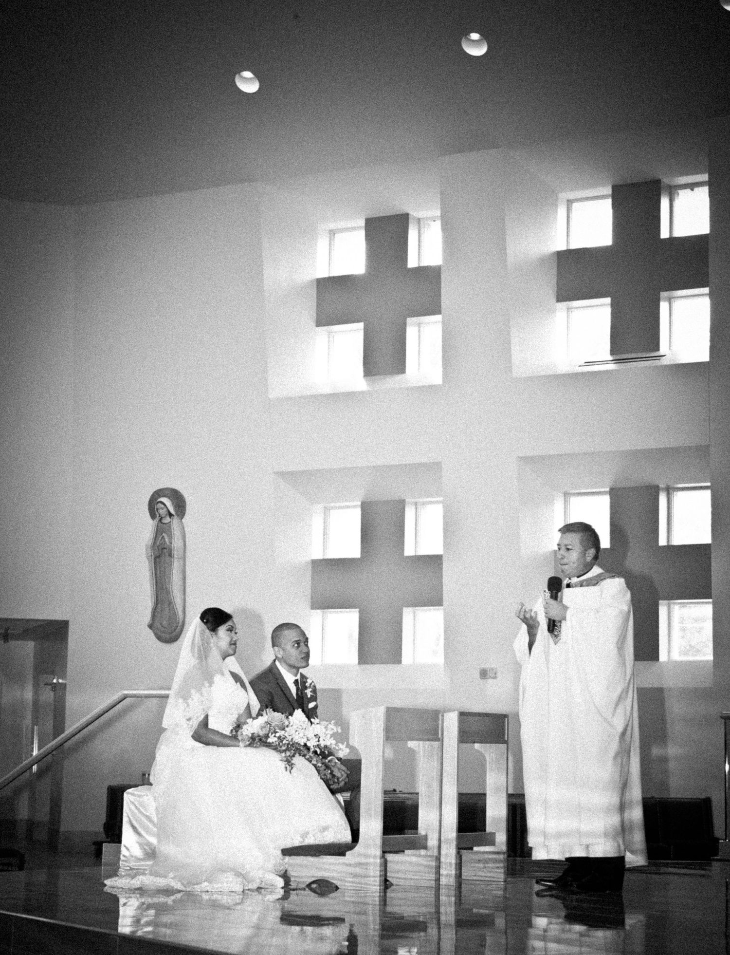 Black and white photo of a wedding ceremony in a modern church with cross windows, bride, groom, and priest at altar.
