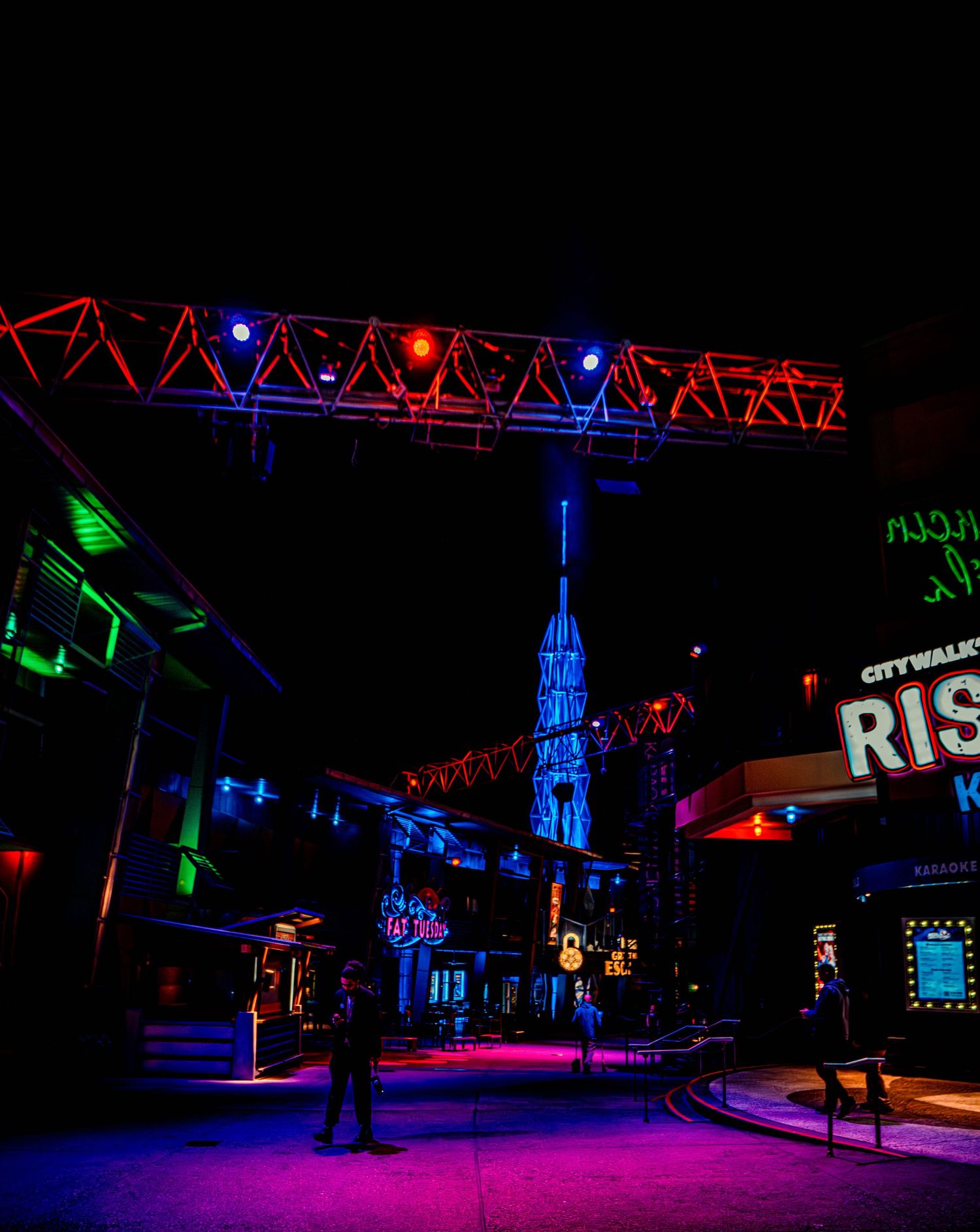 Colorful neon lighting illuminates a dark outdoor stage area with red and blue truss structures creating a dramatic atmosphere.