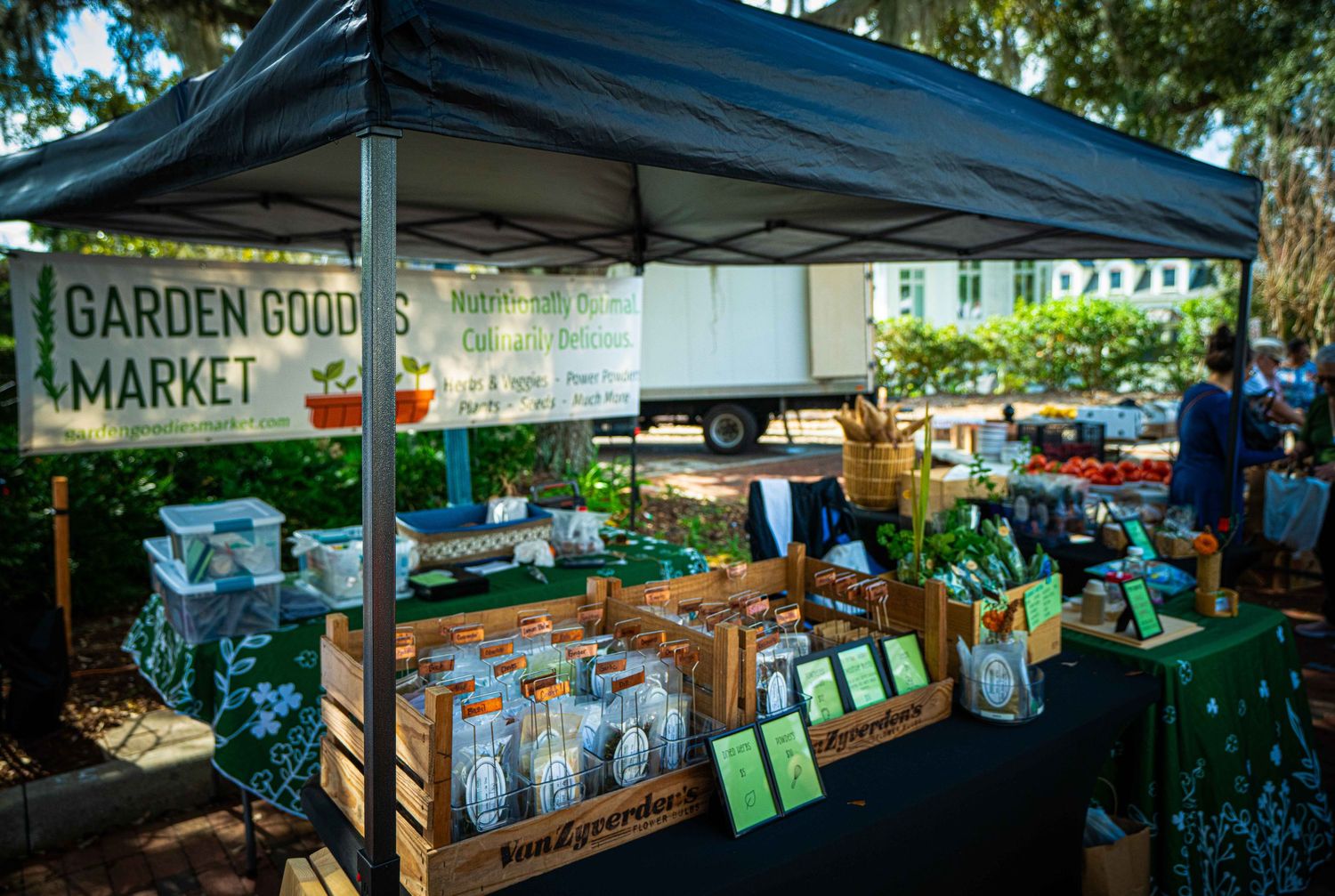 A farmers market stall displays fresh produce and goods under a covered outdoor area on a sunny day.