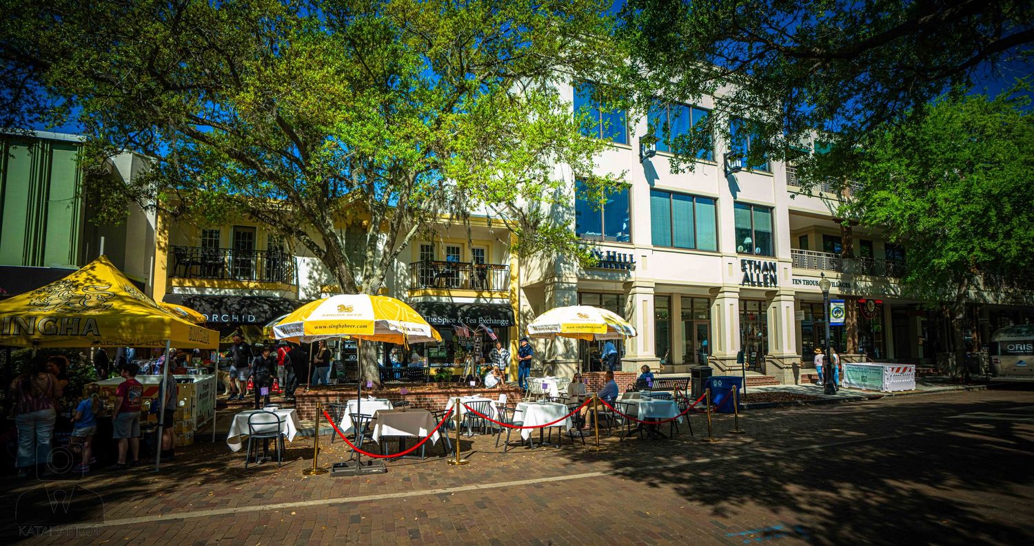 Outdoor cafe seating with yellow umbrellas on a sunny day in front of historic buildings with lush green trees.