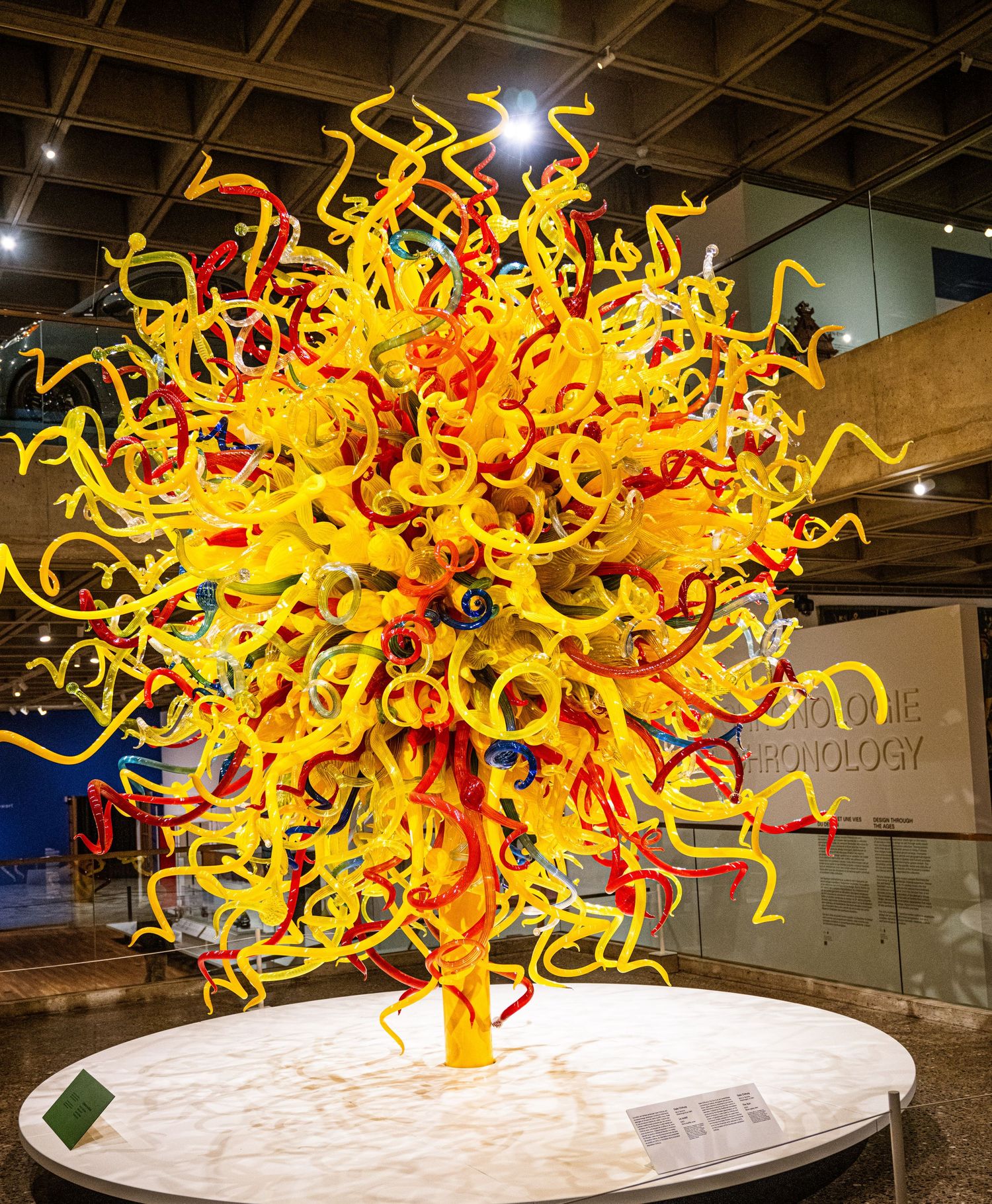A vibrant glass sculpture resembling a sun burst with swirling yellow and red tendrils displayed on a white pedestal.