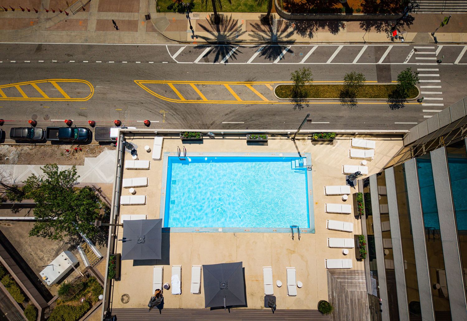 Aerial view of a rooftop swimming pool with lounge chairs on a sunny day, surrounded by city streets.