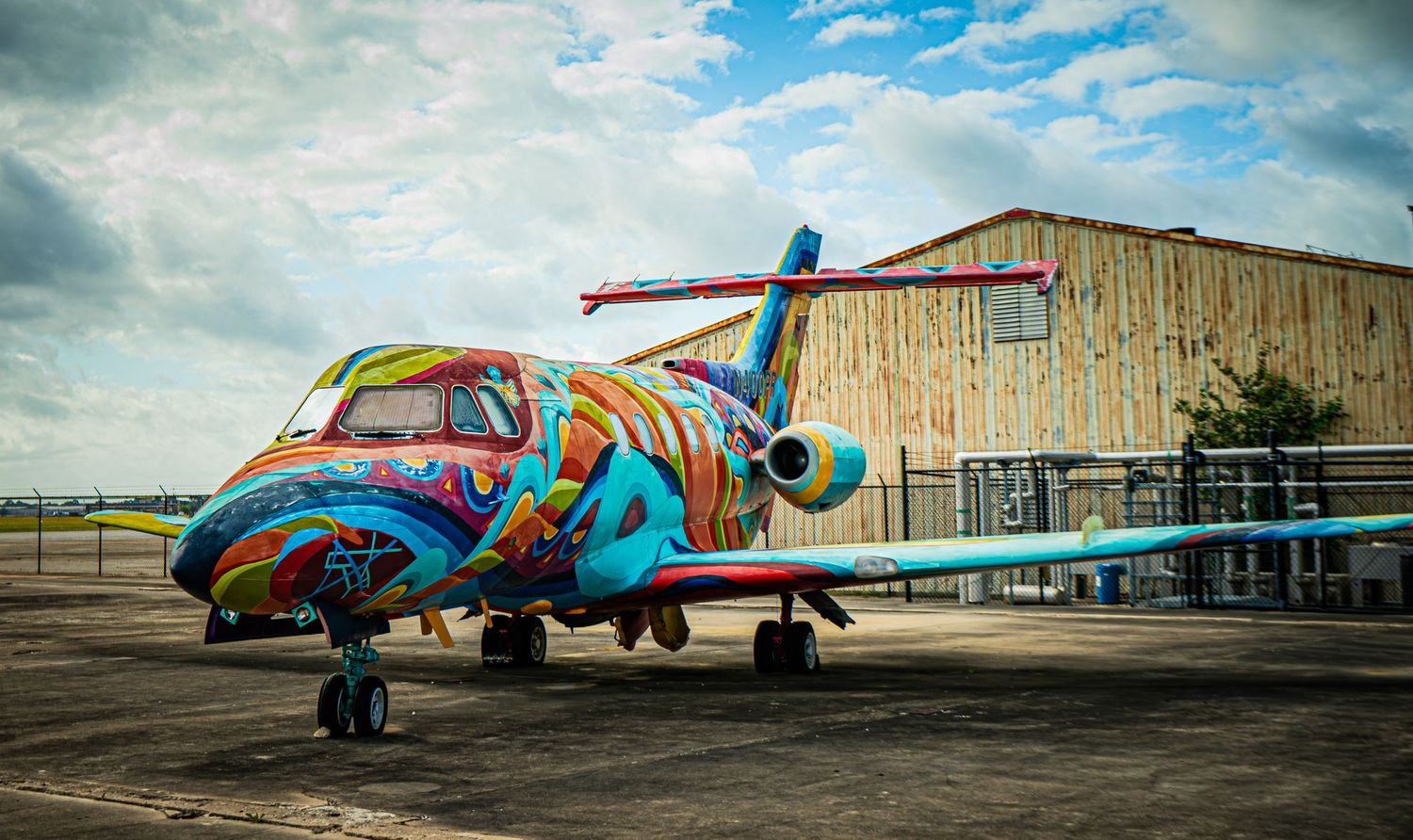 Colorfully painted graffiti art jet airplane parked on tarmac near a wooden hangar under cloudy skies.
