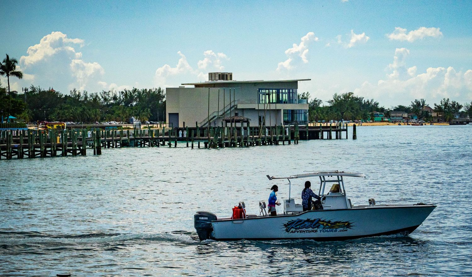 A white center console boat with passengers cruises calm Florida waters near a pier and waterfront building on a sunny day.