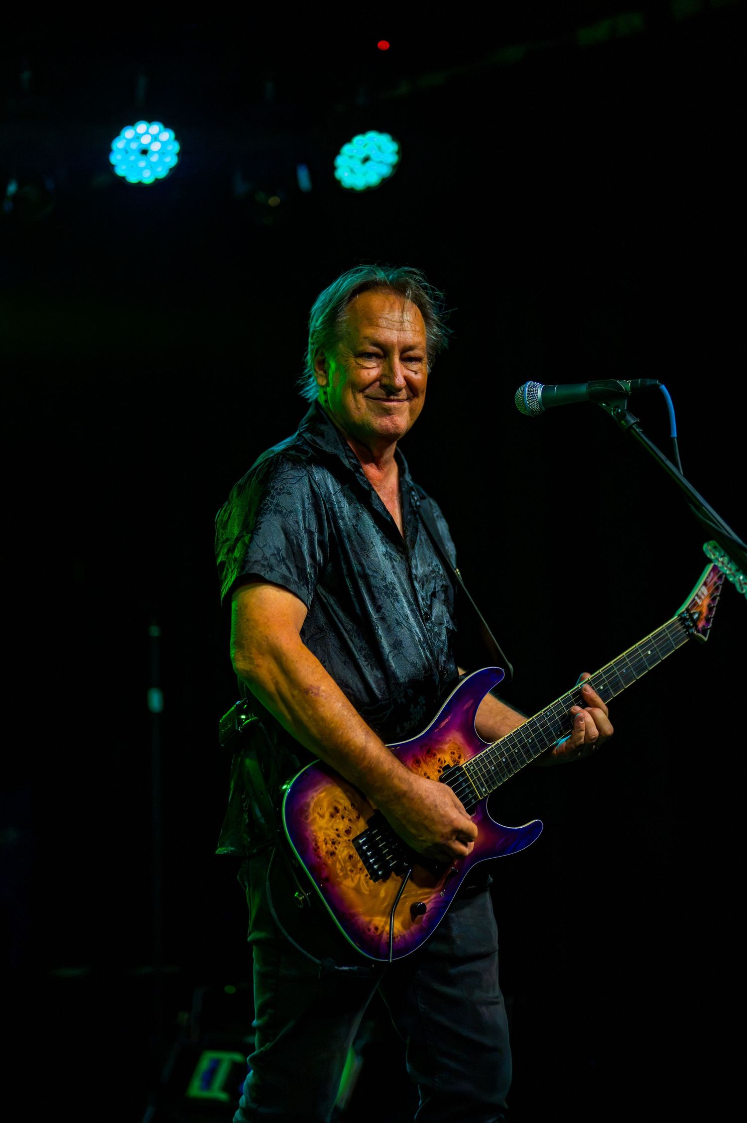 Smiling male guitarist performing on stage with a colorful electric guitar under blue stage lights.
