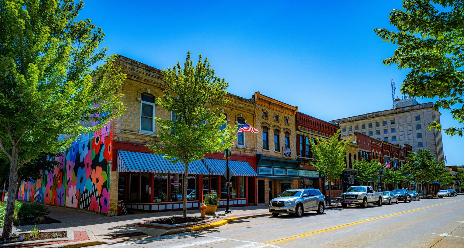 Colorful historic downtown street with brick buildings, parked cars, and trees on a sunny day.