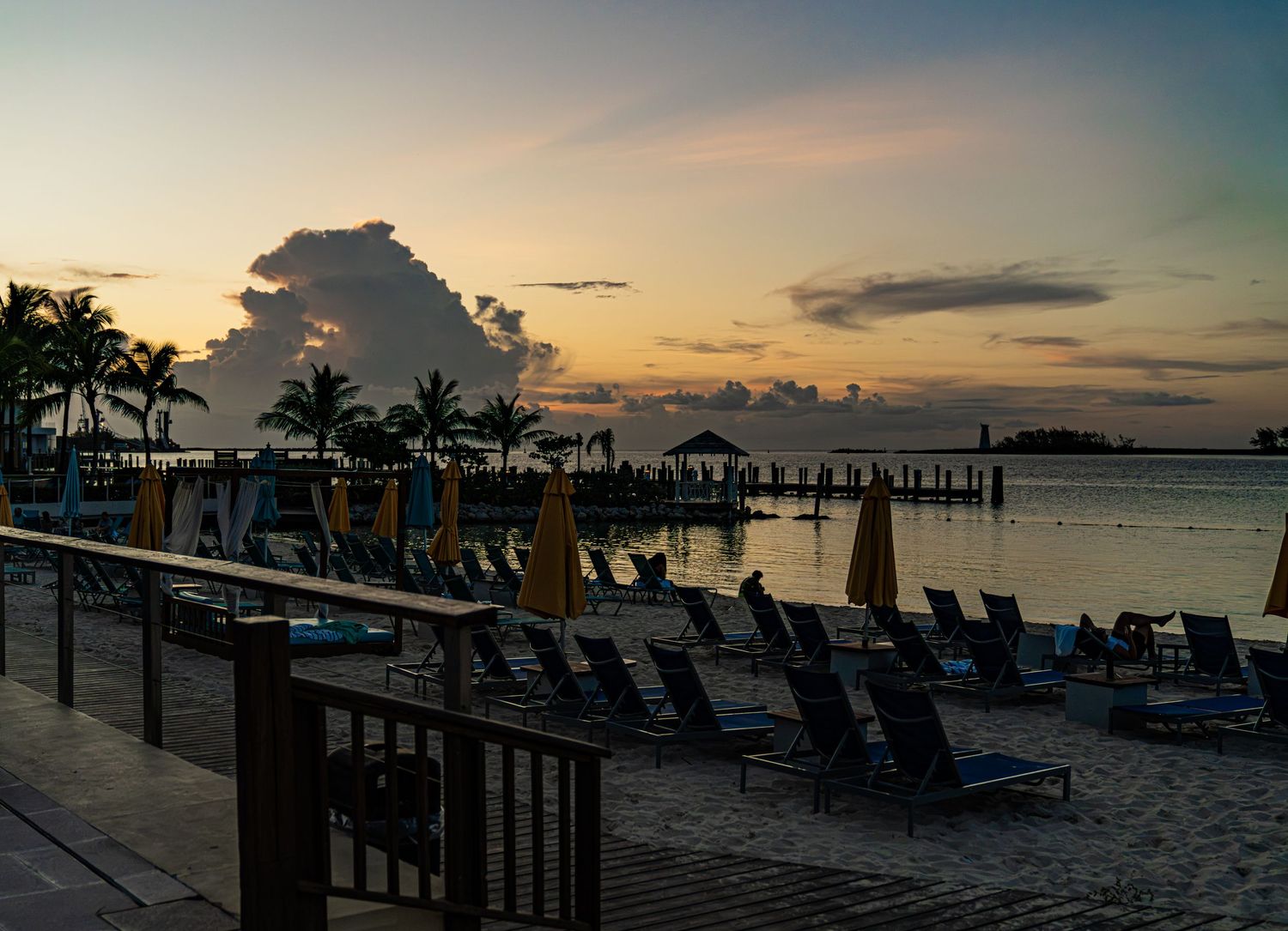 Tropical beach resort at sunset with lounge chairs, palm trees, and a pier overlooking calm waters under a colorful sky.