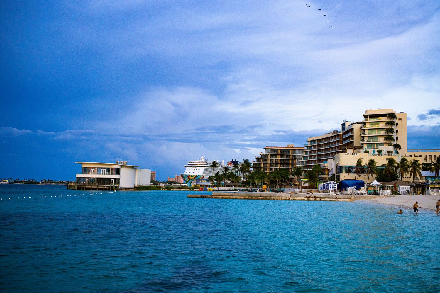 Turquoise Caribbean waters with beachfront hotels and palm trees under a dramatic blue sky.