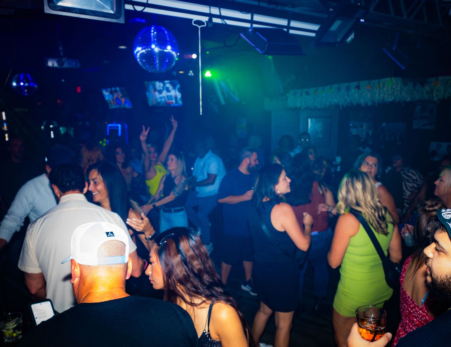 Crowded nightclub dance floor with colorful blue and green lighting as people dance and socialize.