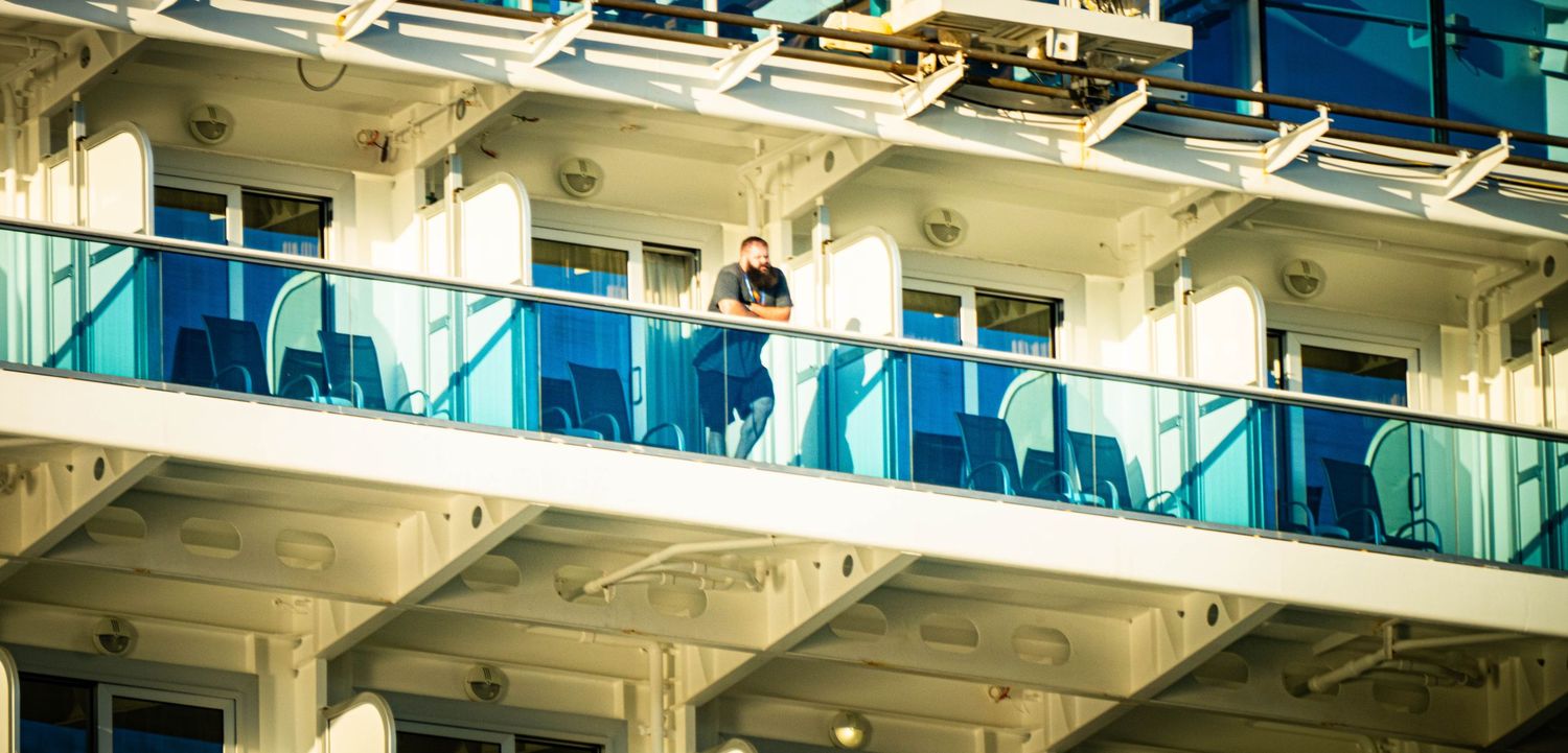 A cruise ship balcony with blue glass railings and white exterior details against a sunny sky.