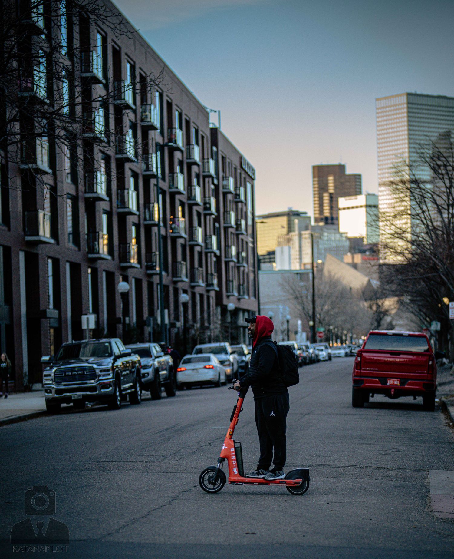 Person riding an electric scooter on an urban street at dusk with modern buildings and parked cars in the background.