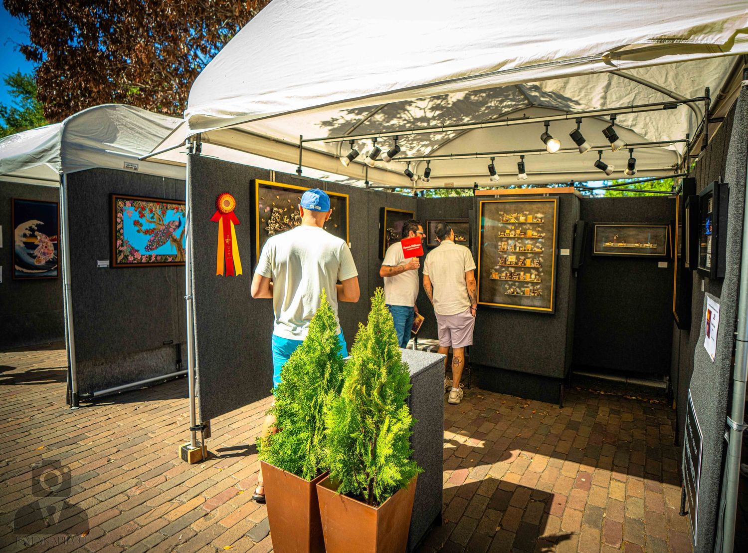 White tent booths display artwork and merchandise at an outdoor art festival under autumn trees.
