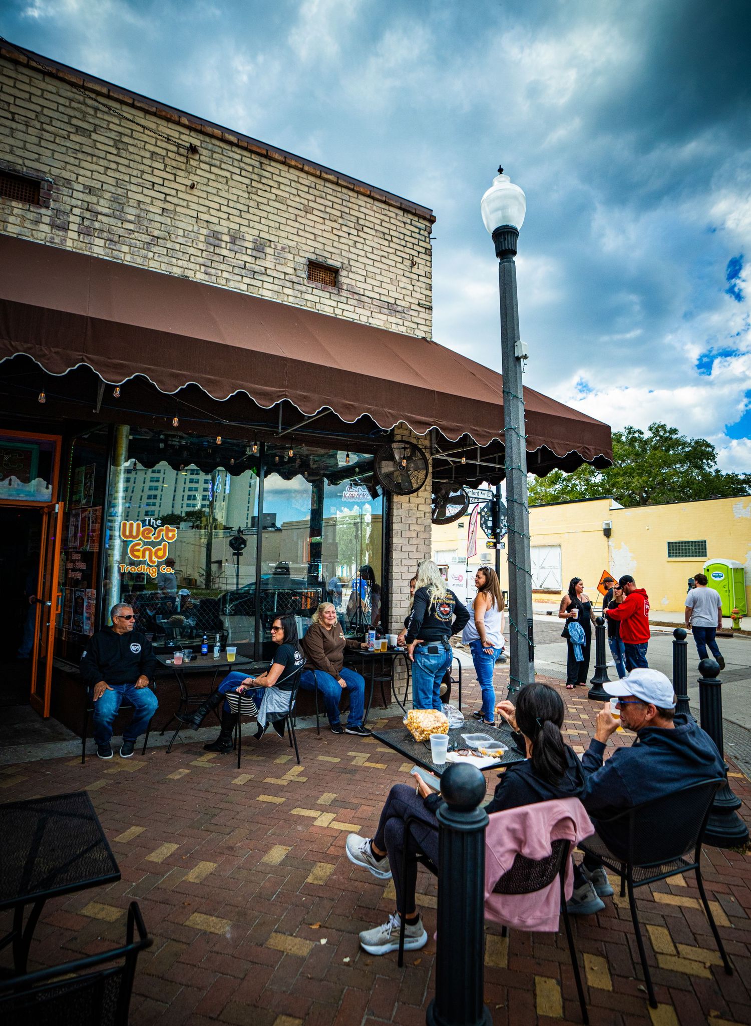 People wait in line outside a brick building on a sunny day with dramatic clouds overhead.