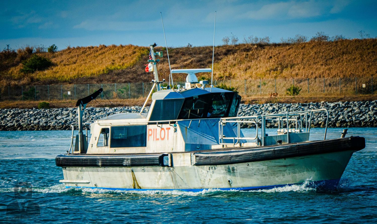 A white and blue patrol boat cruises through coastal waters near rocky shoreline on a sunny day.