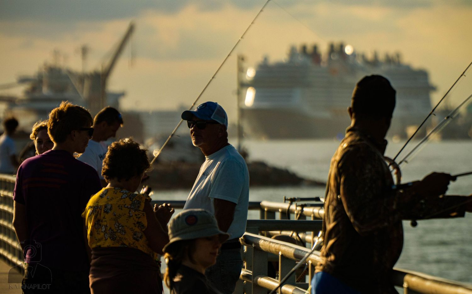 Silhouettes of people fishing from a pier at sunset with boats in the harbor.