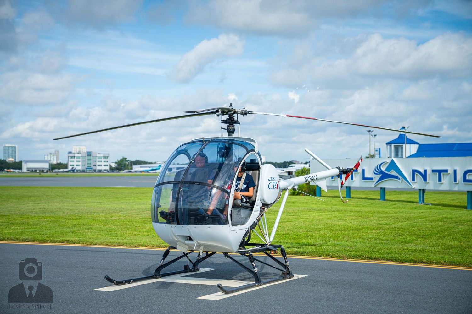 A small white helicopter sits on a grassy airfield tarmac with blue skies and hangars in the background.