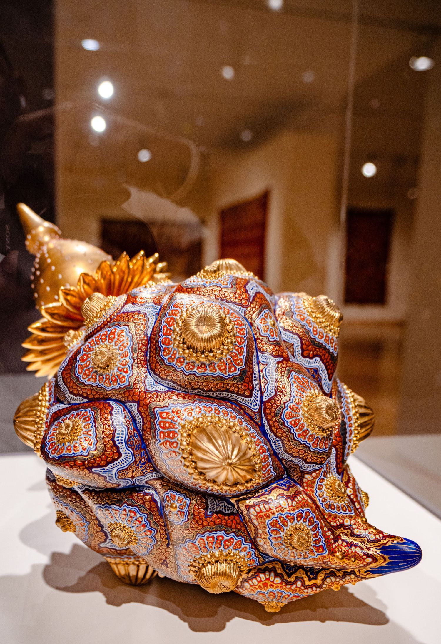 Ornate decorative seashell sculpture with intricate mosaic patterns in blue, orange, and gold displayed in a museum gallery.