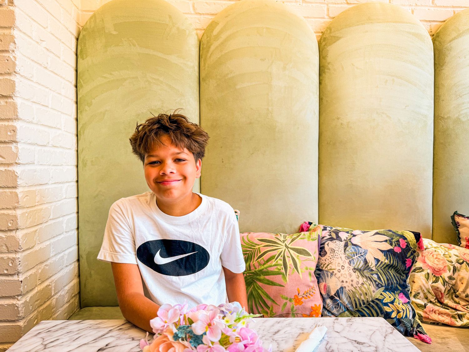 Smiling boy in Nike t-shirt sitting at a cafe booth with floral cushions and green velvet headboard.